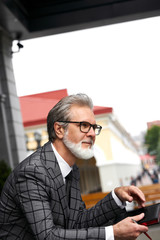 stylish senior man writer thinking abou business plan outdoors, after business meeting, hold notebook in hands and looks away