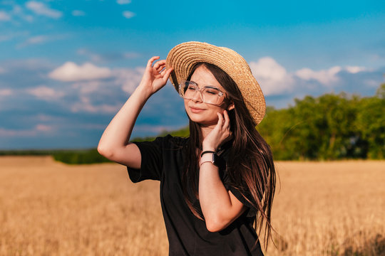 Female outdoors. A young pretty woman in a black undershirt and a straw black hat posing on camera on golden rye meadow background. Concept photo shot in a wheat field late summer