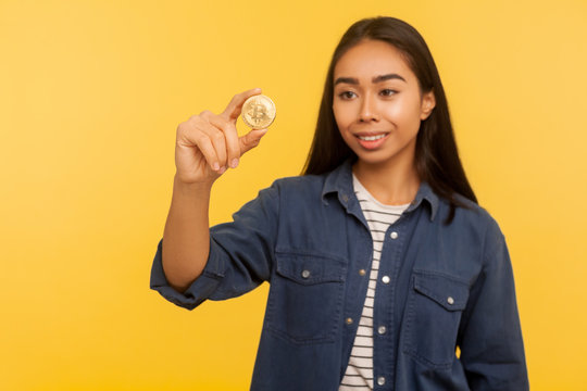 Cryptocurrency btc coin. Portrait of rich happy brunette girl in denim shirt smiling and looking at golden bitcoin, electronic money, digital currency. indoor studio shot isolated on yellow background - Powered by Adobe