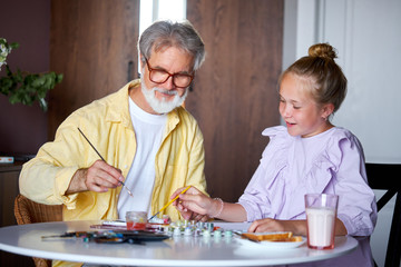 cute little girl and senior granddad painting with colorful paints at home, they spend time together, sit at table, talk and draw