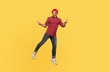 Fototapeta na wymiar Portrait joyful hipster trendy guy levitating, looking at camera with victory peace gesture, hovering in mid-air, smiling and flying up. full length, indoor studio shot isolated on yellow background