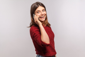 Portrait of cheerful brunette woman in shaggy sweater talking with friend on cell phone, looking away and smiling joyfully, having pleasant conversation. indoor studio shot isolated on gray background
