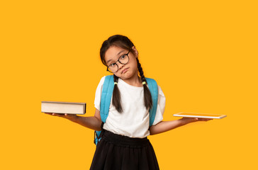 Puzzled Chinese Schoolgirl Comparing Digital Tablet And Book In Studio