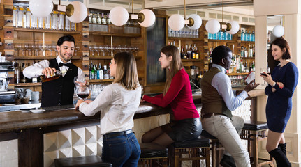 Happy cheerful smiling barman is servicing young people who are relaxing in bar indoor.