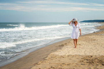 View of a beautiful young woman in white dress and summer hat walking on a sandy beach