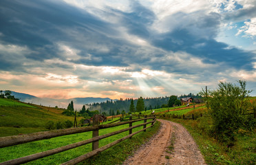 Obraz premium Breathtaking view of sun makes its way through dramatic clouds and dirt road in Voronenko village, Carpathians, Ukraine