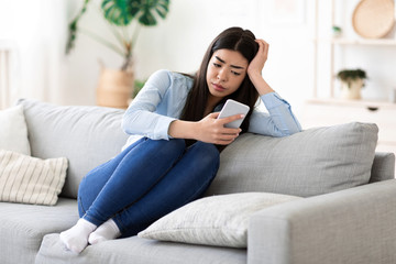 Obraz premium Bored At Home. Weary Asian Girl Sitting With Smartphone On Sofa