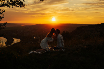 Silhouettes of loving couple hugging and kissing while sitting on blanket at summer picnic at gorgeous sunset. Summer  time, mountains and sunset in background. Copy space.