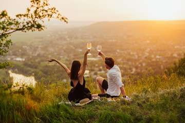 Couple sitting at summer picnic with hands up and watching sunset at a mountain, while making a toast with wine during golden hour.