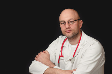 Portrait of a bald doctor in white uniform and red stethoscope on a black background. Hands crossed.