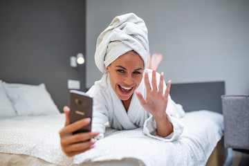 Smiling young woman lying on bed in bathrobe with her mobile phone taking a selfie.Beautiful woman relaxing on the bed after bath and looking at the phone camera taking a selfie