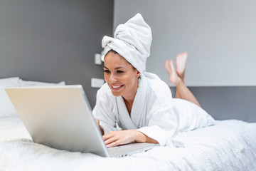 Young beautiful woman in towel and bathrobe sitting on bed at home and working on her laptop computer. beautiful young woman wearing a white bathrobe, with a towel on her head in bed at home