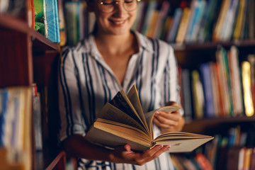 Young smiling attractive college girl leaning on book shelves in library and searching for material...