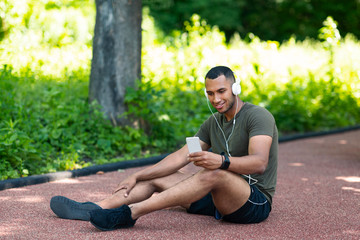 Relaxed black man sitting on jogging track and listening to music from smartphone at park