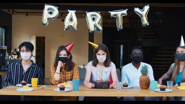 Sad weird party celebration with safety measures. Upset multiethnic friends in masks sit at table together slow motion.