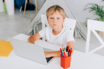 high angle view of boy in white t-shirt using laptop while sitting at table near pen holder and kids wigwam on background © LIGHTFIELD STUDIOS