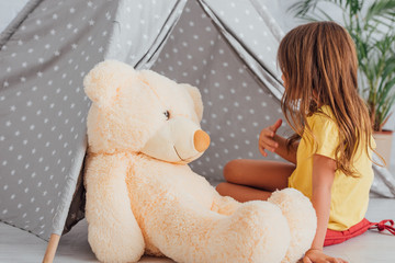 girl in yellow t-shirt talking to teddy bear while playing near kids wigwam © LIGHTFIELD STUDIOS