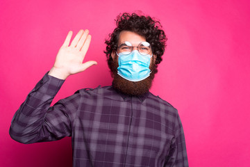 Photo of happy young man with curly hair wearing medicinal mask and saying Hello.