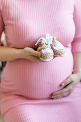 Close-up shot of the belly of a 30 weeks pregnant woman wearing a light pink bodycon dress in a light room. A woman holds booties for a baby in her hands. Pregnancy planning and preparation for child