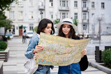 Two young brunette girls, wearing casual jeans clothes, standing in front of old historical building, holding travel map, checking route. City sightseeing tour.Tourism lifestyle.