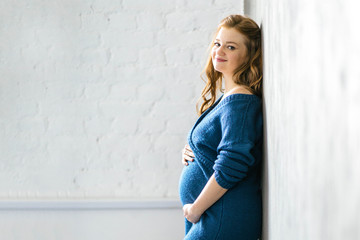 30 weeks pregnant woman wearing a blue bodycon dress stays by the white wall. Going on maternity leave, planning pregnancy, preparing for childbirth