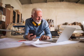 Elderly engineer holding cellphone and looking at the laptop - obrazy, fototapety, plakaty