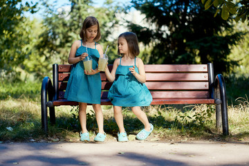 two cute little girls in dresses sit on a bench and drink lemonade in the summer