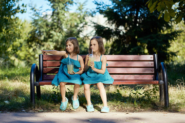 two cute little girls in dresses sit on a bench and drink lemonade in the summer