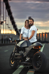 Naklejka premium Young sexy woman hugging cute man in stylish black leather jacket, sitting on sports motorcycle on the bridge in the city on sunset and kissing