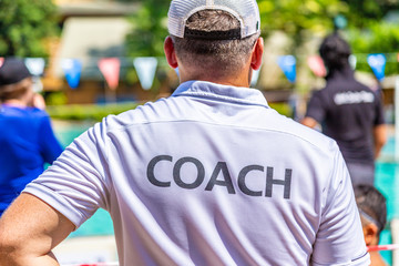 Male swimming coach in white COACH shirt standing in the sun watching his swimmers competing in the pool