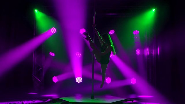 Sexy brunette in a swimsuit with rhinestones and high heels makes rotational movements on the pole. Performing in a nightclub in the twilight and bright red spotlights. Silhouette.