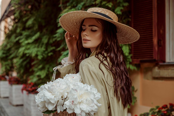 Young elegant brunette woman with long waved hair wearing wide brim straw hat, holding white peony...