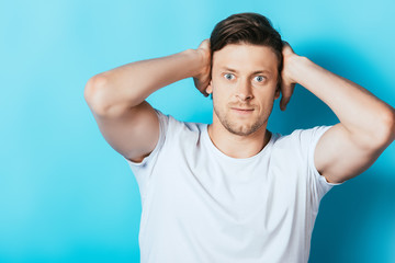 Fototapeta na wymiar Angry man in white t-shirt covering ears with hands on blue background