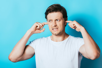 Aggressive man in white t-shirt covering ears with fingers on blue background