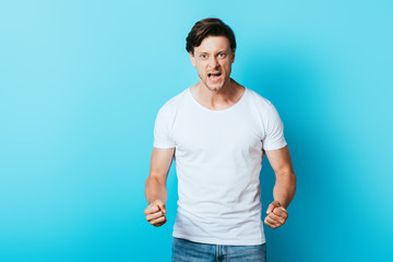 Irrated man in white t-shirt with hands in fists looking at camera on blue background