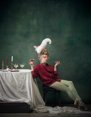 Enjoy music with wine glass. Young woman as Marie Antoinette on dark green background. Retro style, comparison of eras concept. Beautiful female model like classic historical character, old-fashioned.