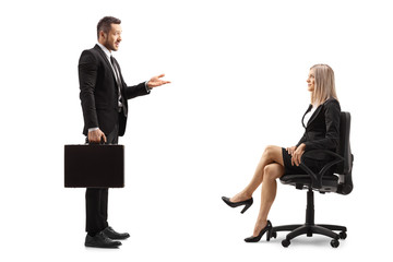 Full length profile shot of a man in a formal black suit talking to a businesswoman sitting in a...