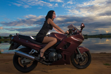 Fototapeta na wymiar a beautiful girl in a short black dress is sitting on a sports motorcycle against the background of a lake at sunset. colorful sky. sandy beach.