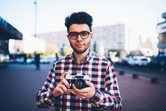 Portrait of skilled male photographer using vintage camera for taking photos outdoors in urban settings, handsome hipster guy spending free time on hobby holding old equipment for making pictures
