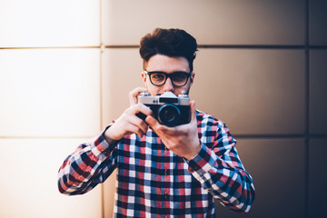 Young man in eyeglasses holding vintage camera making settings before taking picture, skilled photographer in cool outfit using old equipment talking pictures focusing lens standing outdoors