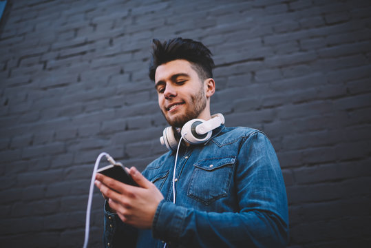 Positive male blogger share photos in networks while resting outdoors standing on wall background, young meloman making settings in playlist before listening music via modern headphones and mobile