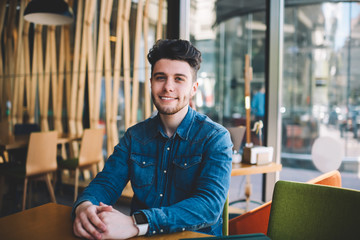 Portrait of cheerful bearded hipster guy in casual denim shirt sitting at table in cafeteria on free time,successful happy young man visit restaurant on leisure in good mood looking at camera