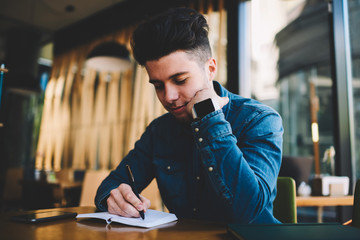 Hipster guy concentrated on planning schedule writing in notepad while sitting in cafe interior,...