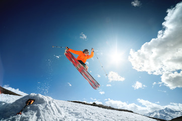 A young stylish man in sunglasses and a cap performs a trick in jumping with a kicker of snow against the blue sky and the sun on a sunny day. The concept of park skiing in winter or spring © yanik88