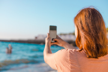 Happy woman on vacation photographing with a camera on the beach of the sea by smartphone, enjoys leisure and summer vacation