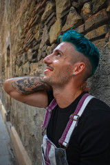Young man wearing overalls, with blue hair, leaning against an old brick wall