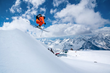 Naklejka na ściany i meble A male skier in an orange suit flies in the air after jumping from a snow sweep high in the Caucasian mountains on a sunny day amid snow-capped mountains of blue sky and white clouds