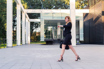 Fototapeta na wymiar A pregnant businesswoman walking in front of a modern building and looking on her wristwatch.