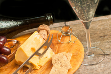 top view, close distance of a glass of champaign and a rare wood plater of wedge of Swiss cheese, cheese knife, crackers , cork and wire retention lock with the neck of empty bottle, on wind table top