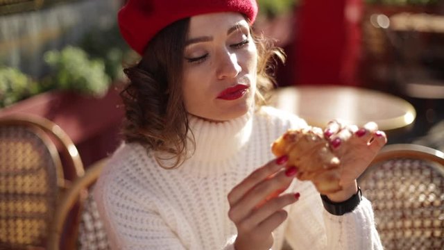 Beautiful woman in red beret sitting outside and eating croissant . French style